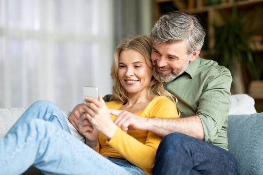Happy Middle Aged Couple Relaxing With Smartphone On Couch At Home