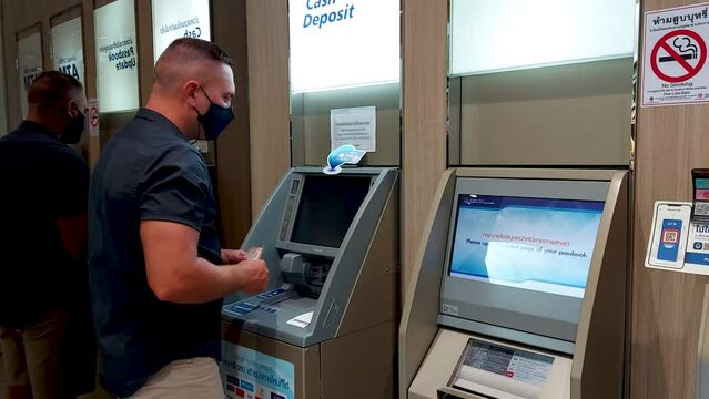 A European man withdraws cash from a Thai ATM, a masked man makes transactions at a Thai bank ATM