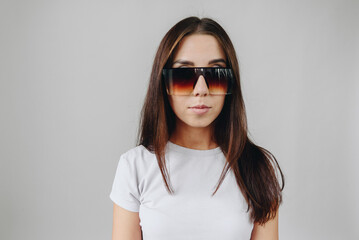 A brunette girl in a white T-shirt on a white background is standing in huge sunglasses
