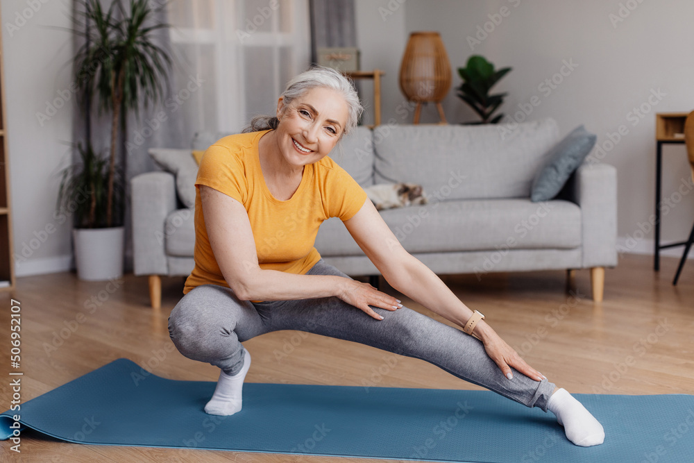 Wall mural Active senior woman doing stretching exercises during her home workout, training, keeping fit and healthy