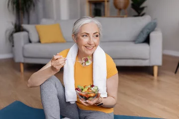 Fotobehang Healthy lifestyle concept. Happy senior woman eating fresh vegetable salad, sitting on yoga mat after home workout © Prostock-studio