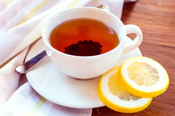 A white cup of tea, two slices of lemon on a saucer are against the background of a wooden table.