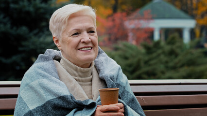 Happy mature single middle aged woman covered with warm plaid drinking hot coffee disposable cup healthy cheerful senior pensioner enjoying rest outdoors smiling old lady sitting on autumn park bench