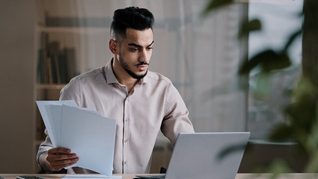 Serious arabian young business guy male professional worker employee type on computer keyboard copy paper text at electronic form focused hispanic man compare documents check online financial report