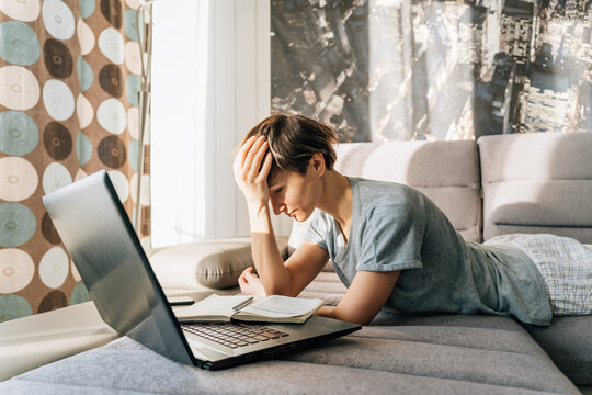 Woman annoyedly holds on to her head lying on the couch near laptop