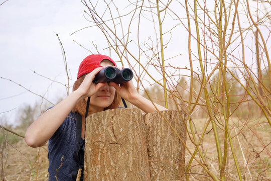 Young girl in spring in thickets of grass and bushes watches birds and wildlife through binoculars. Birdwatching