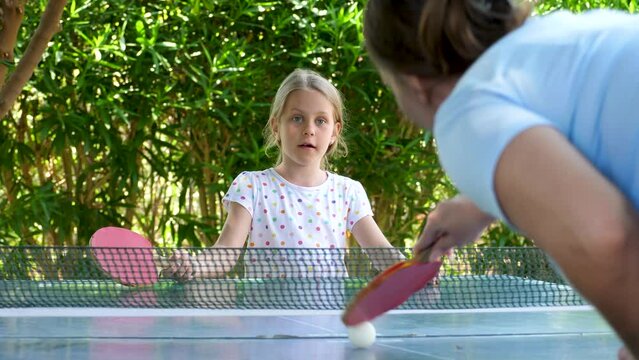 Playful youth girl play table tennis in garden. Child player hit lightweight ping-pong ball back and forth across hard table divided by tennis net use small red rackets. 