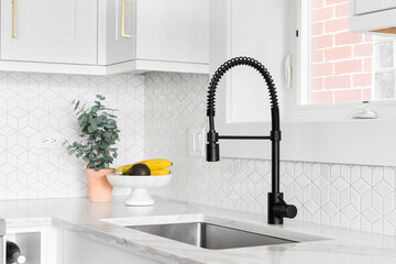 A farmhouse kitchen sink detail shot with a black faucet, mosaic tile backsplash, marble countertops, and white cabinets.