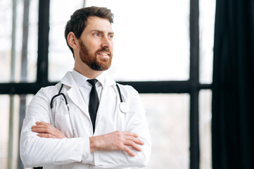Caucasian proud handsome male doctor, therapist, surgeon, cardiologist in a medical uniform and stethoscope, standing in a hospital, looking to the side with arms crossed, smiling, dreaming, thinking