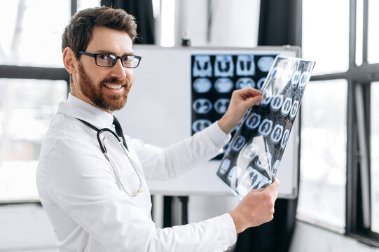 Handsome positive male caucasian doctor holding x-ray at his cabinet, male therapist in medical uniform and glasses learning patient's x-ray, standing in his medical office, looks at camera, smile