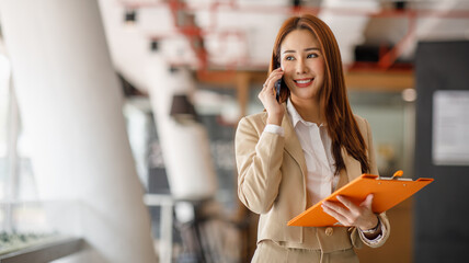 Charming Asian businesswoman manager with a smile standing holding document and talking on mobile...