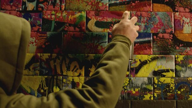 Back view young boy painting colorful graffiti on tiles or street wall with color aerosol . Rear view man in hoodie spraying with  color bottle . Modern , abstract street art . Slow motion . 