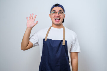 Young asian barista man wearing apron greeting client waving to say hello or hi to customer