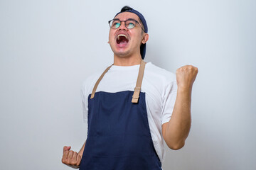 Asian barista man wearing apron standing very happy and excited doing winner gesture with arms raised, smiling and screaming for success. Celebration concept.