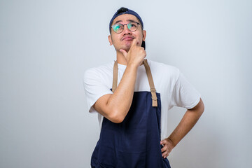 Portrait of attractive Asian barista man in blue apron thinking about question with hand on chin