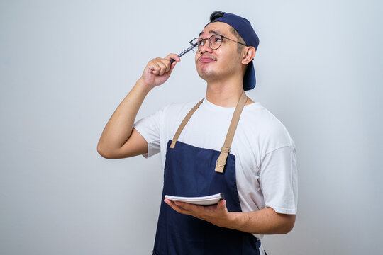 Asian barista man in blue apron taking order, writing on menu book list, thinking with pen on head