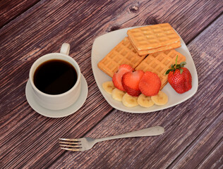 Fresh Viennese waffles with strawberries and bananas on a platter, next to a cup of black coffee on a wooden table.