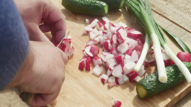 a hand cuts a radish with a knife on a cutting board next to leeks and cucumbers