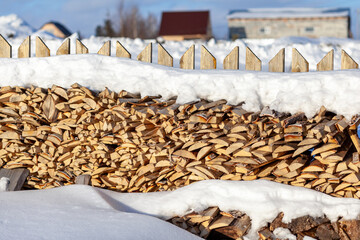 Firewood stacked in a woodpile along a fence in winter under snow against a blue sky. Preparation of firewood for a bath and heating a house