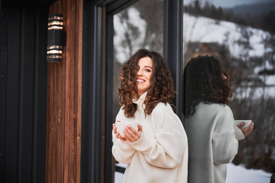 Curly young woman resting on terrace of modern barn house in the mountains. Portrait of happy female tourist holding cup of tea, enjoying in new cottage in winter.