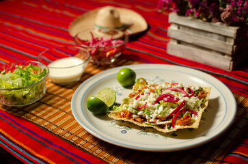 Tostada de tinga de pollo con queso, crema, cebollas con habanero, limones y lechuga; colocado sobre un rebozo rojo. Sombrero de fondo.
