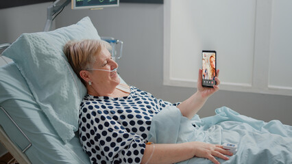 Senior patient talking to family on video call in hospital ward bed. Woman with sickness using...