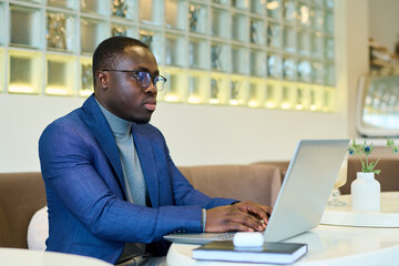 African freelancer in eyeglasses sitting at table and typing on laptop, he working online at cafe