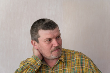Obraz premium Portrait of a grown man in a plaid shirt against a beige background. A man with a short haircut and graying hair has his arm around his neck from behind. Front view. Look past the camera. Indoors.