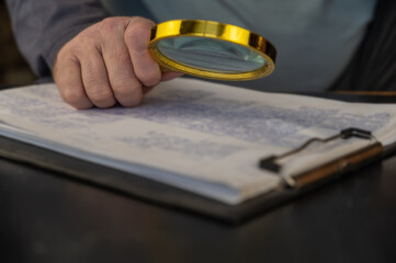 Handholds a gold-rimmed magnifying glass atop a clipboard. Adult male examining documents with handwritten text. Close-up. Selective focus.