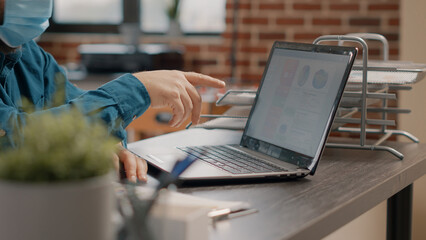 Close up of employee working with laptop and charts files, wearing face mask at desk. Business man using technology and rate information to design project and presentation during pandemic