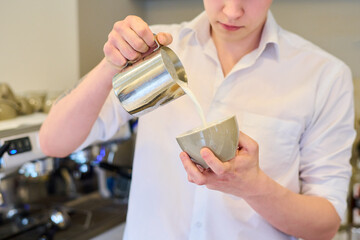 Close-up of young barista pouring milk in cup to make fresh coffee in cafe