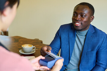 Smiling African man sitting at table with coffee and paying for his order to waitress with credit card
