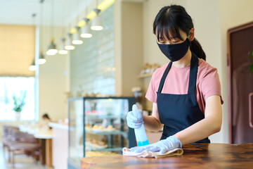 Young waitress in uniform and protective mask cleaning table in cafe with spray of disinfection...