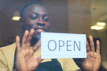 Smiling African man hanging open sign on window of his cafe before the beginning of work day