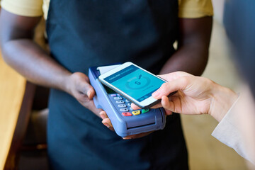 Close-up of customer putting mobile phone on terminal while waiter holding it and paying for her...