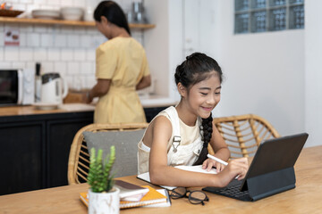 Portrait of little girl using laptop during online class with tutor or teacher while sitting at desk in cozy kitchen interior with mother in background