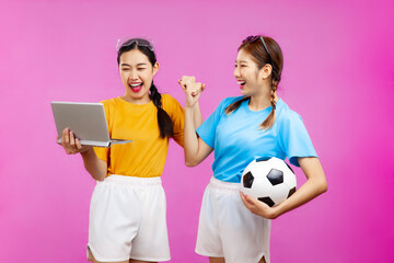 Two Cheerful Asian woman over isolated pink background holding a soccer ball and laptop, whatching football match, sport concept