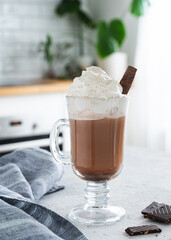 Hot chocolate with whipped cream and chocolate pieces in a tall glass with a cinnamon stick on the background of a white kitchen in the  early morning. Front view and close up