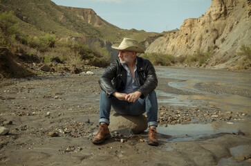 Adult man in cowboy hat sitting on rock along river flows in desert. Almeria, Spain