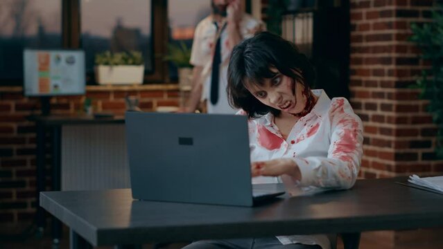 Creepy looking office zombie woman working on modern laptop while coworker talking on landline. Bizarre scary brain-eating monsters inside company workspace using portable computer and telephone.