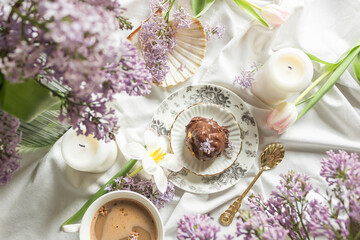 Top view of breakfast in bed with antique tableware, cup of coffee and chocolate profiterole, decor of spring flowers.