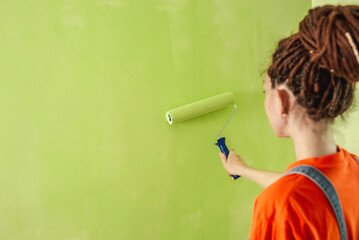 Woman with dreadlocks, in an orange t-shirt and denim jumpsuit is painting the wall with a roller in green color. Concept of repair, renovation of the new apartment. Copy space