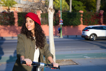 Beautiful young latin woman with curly brown hair wearing red cap and dressed in casual clothes is sightseeing in europe visiting the city on scooter. Tourism concept.