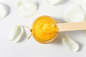 Glass jar with orange sugar body scrub on a white table with flower petals.