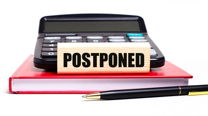 A burgundy notebook on a white background. On it is a black calculator, a pen and a wooden block with a POSTPONED text. Business concept