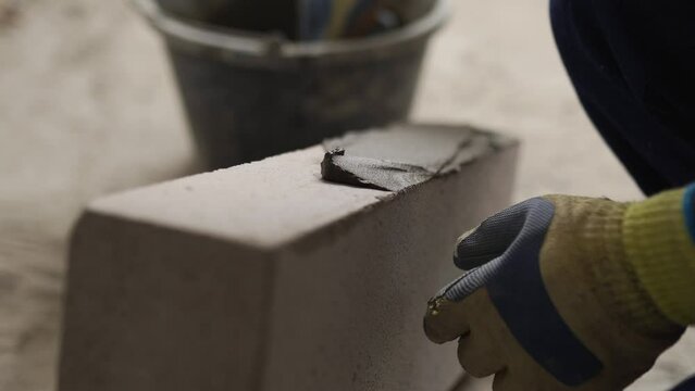 Bricklayer applies adhesive glue on autoclaved aerated concrete blocks with notched trowel and spatula. Brickwork worker contractor puts mortar on foam concrete doing masonry on construction site.