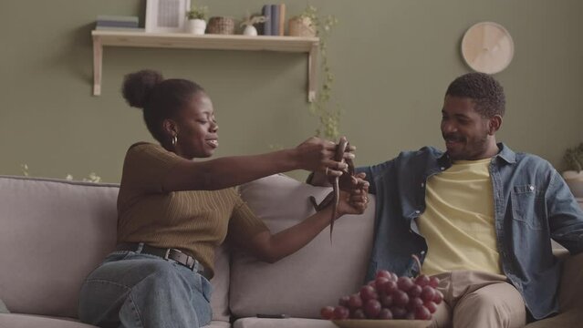Medium slowmo of young African American couple with pet snake sitting on sofa in olive green walled living room with many houseplants