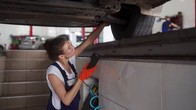 Female mechanic inspecting car suspension on hydraulic wheel play detector with remote control. Strong woman checks for wear in running gear and steering components. Service operator in overalls.