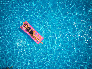 Fit pretty girl in bikini chilling on inflatable pink mattress in swimming pool. Slim hot woman in swimwear tanning. Female relaxing on float in blue water at luxury resort. Aerial, view from above.