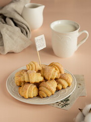 Pastry rolls. Mini cottage cheese croissants. Bagels with sugar on the milky color ceramic plate. Mug with milk, milk jug an beige linen kitchen towel